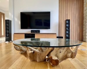 Modern living room featuring a large glass coffee table with a natural wood base, flanked by home theatre speakers, with a television mounted on the wall.