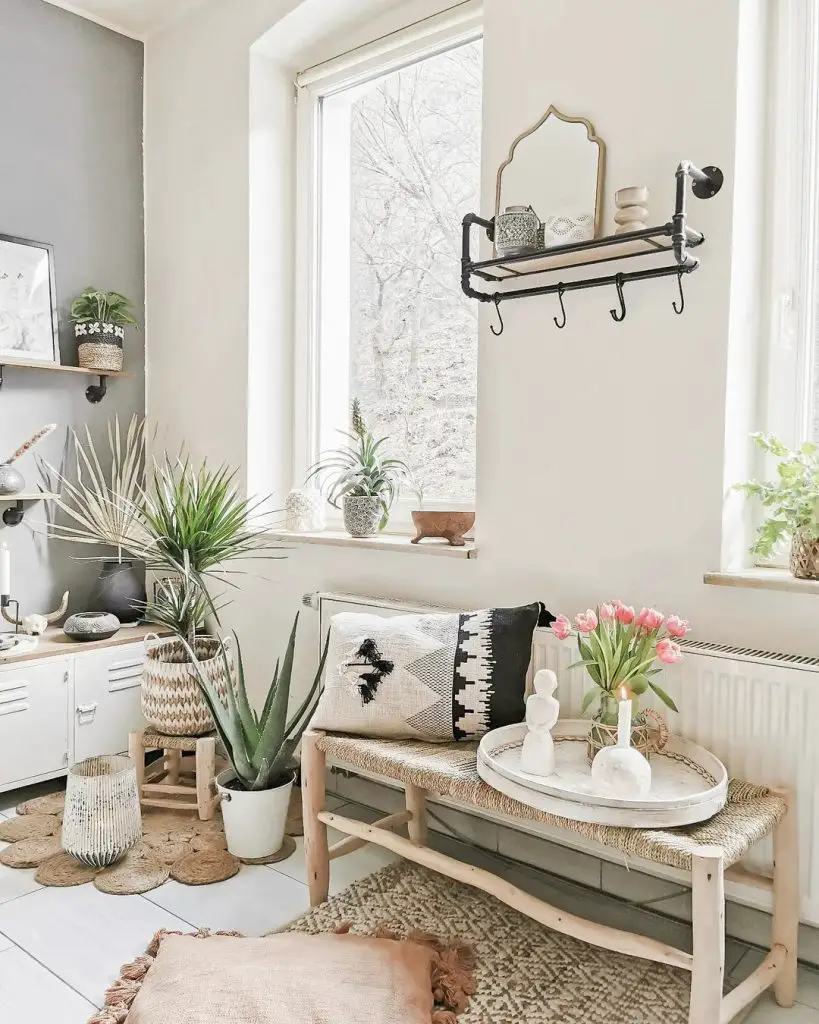 A cozy corner of a room featuring a white sofa with decorative pillows, a small wooden bench, houseplants, and a round mirror above a rustic shelf adorned with boho decor.