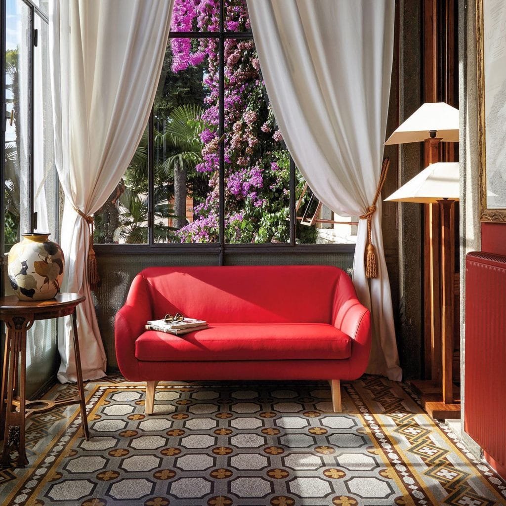 A bright living room featuring a vibrant red sofa, decorative curtains, and a large window showcasing purple flowers outside.