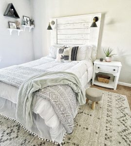 A cozy bedroom featuring a white bed with modern farmhouse bedding, a small wooden stool, a white nightstand, and decorative wall items in a light, neutral-toned room.