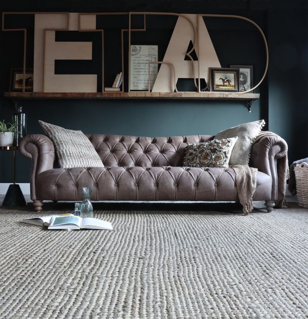 A luxurious brown leather chesterfield sofa in a room with dark blue walls and a large "TETRAD" sign above, exemplifying the principle of maximizing negative space for a calming and uncluttered home. A book lies open on the textured carpet.
