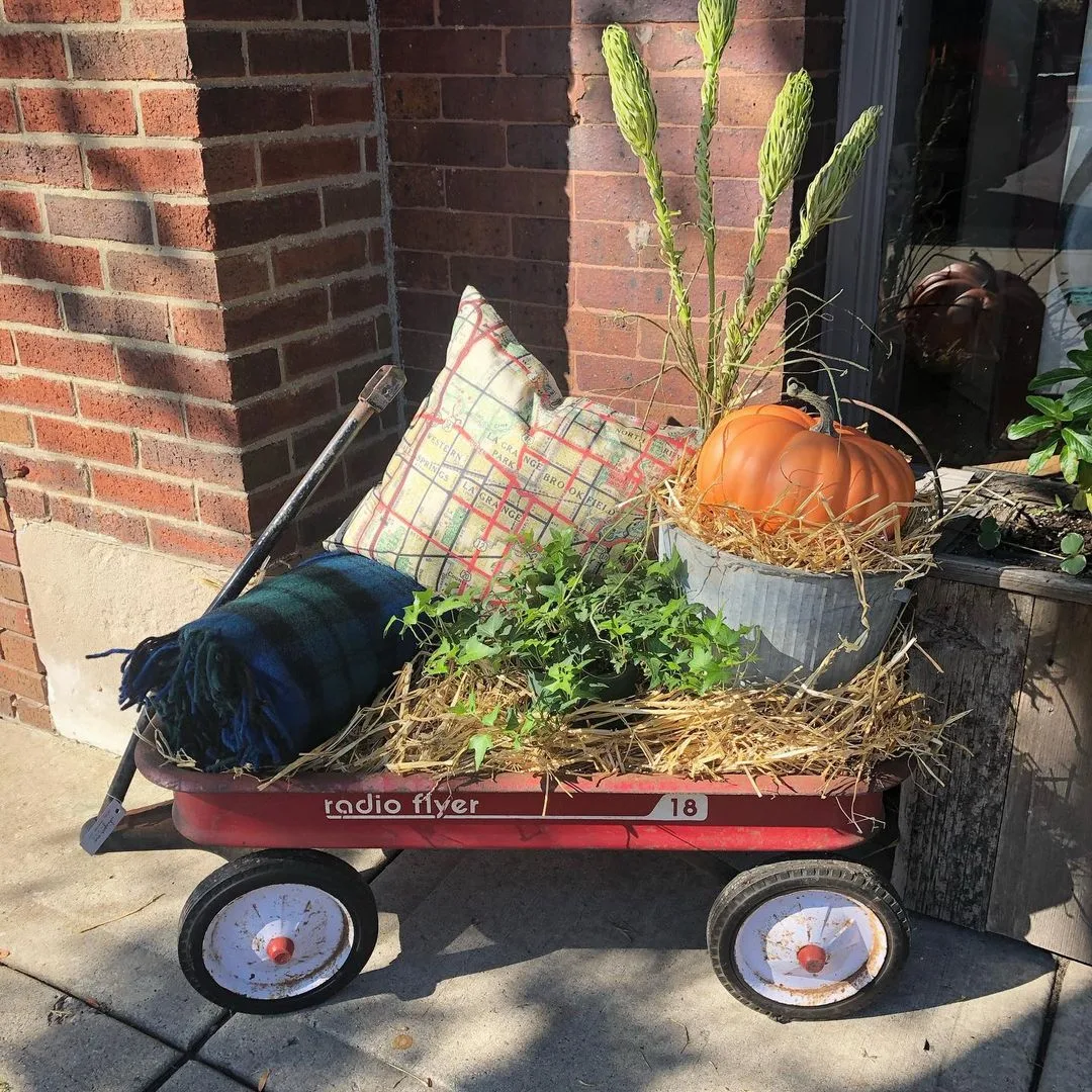A red Radio Flyer wagon filled with a pumpkin, plants, a plaid pillow, a blanket, and straw creates one of the unique vignettes by a brick wall to decorate an amazing front porch.