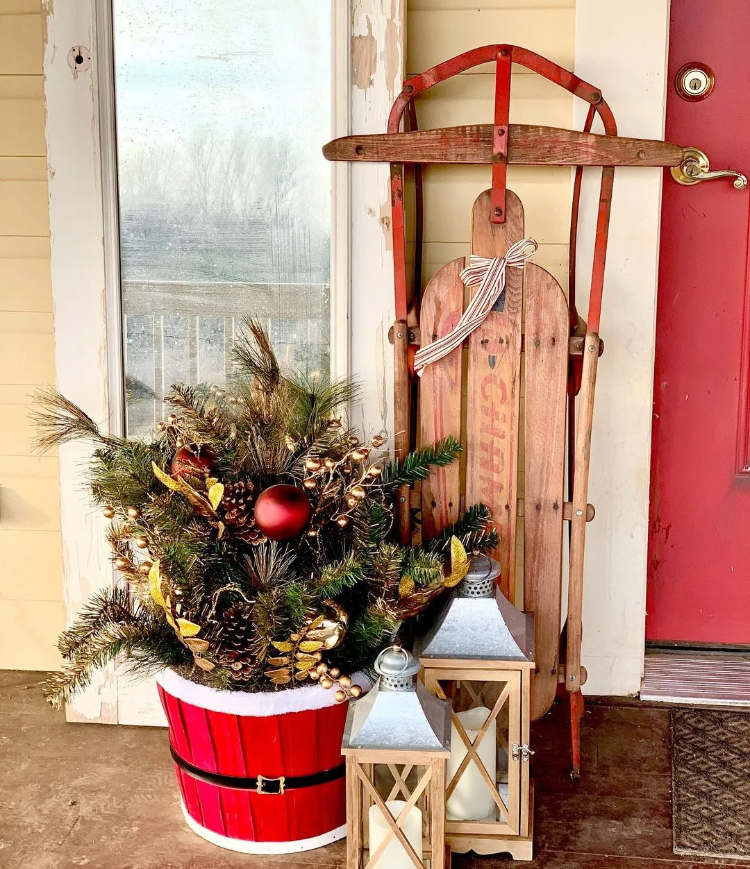 Vintage wooden sled and ski propped against a red door, next to a red bucket with a unique Christmas pine and two white lanterns on an amazing front porch.
