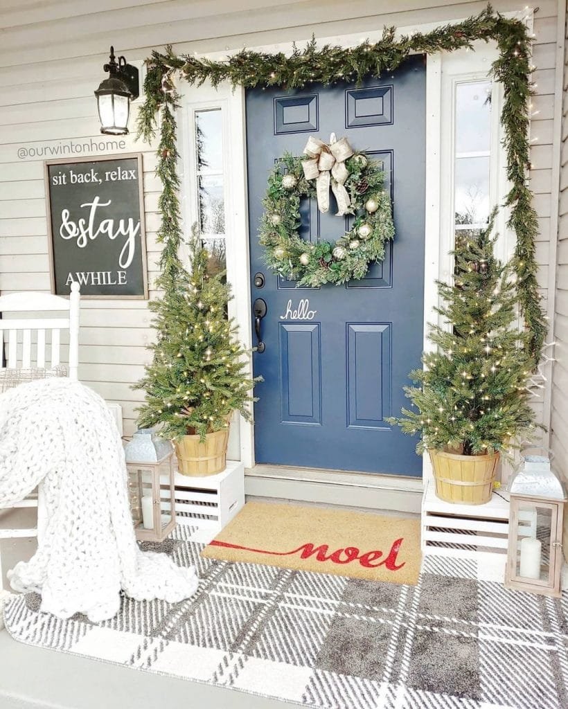 A front porch decorated for the holidays with a blue door, wreath, two small Christmas trees, lanterns, a plaid rug, and a cozy blanket draped over a white chair, perfect inspiration for Christmas Front Porch Decor Ideas.