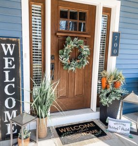 a natural wooden front porch door with a door wreath, a welcome sign, a welcome door mat, and potted flowers ready for the Spring season