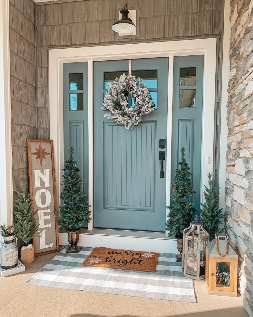 A festive front porch painted pale blue, adorned with a silver wreath, flanked by small Christmas trees and holiday decor including a "NOEL" sign.