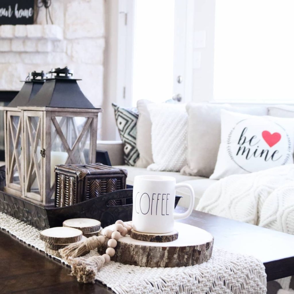 A cozy living room setup featuring a coffee mug on a wood coaster, decorative lanterns, and a throw pillow with "be mine" text and a heart. White sofa with patterned pillows in the background adds to the rustic charm, complemented by wood stumps used as table decor.