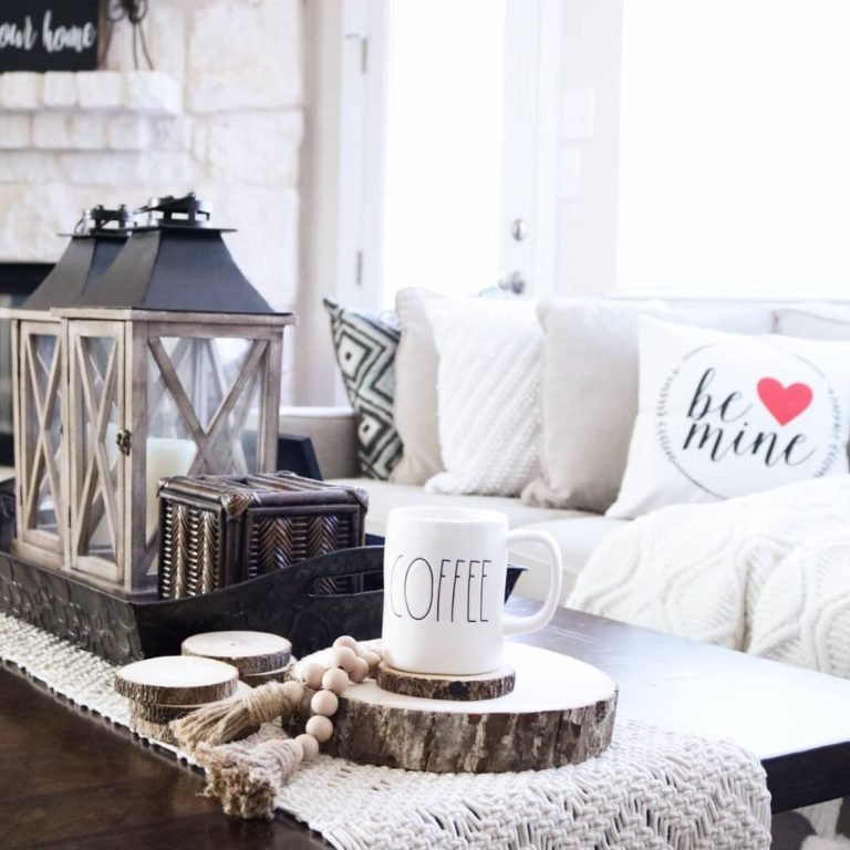 A cozy living room setup featuring a coffee mug on a wood coaster, decorative lanterns, and a throw pillow with "be mine" text and a heart. White sofa with patterned pillows in the background adds to the rustic charm, complemented by wood stumps used as table decor.