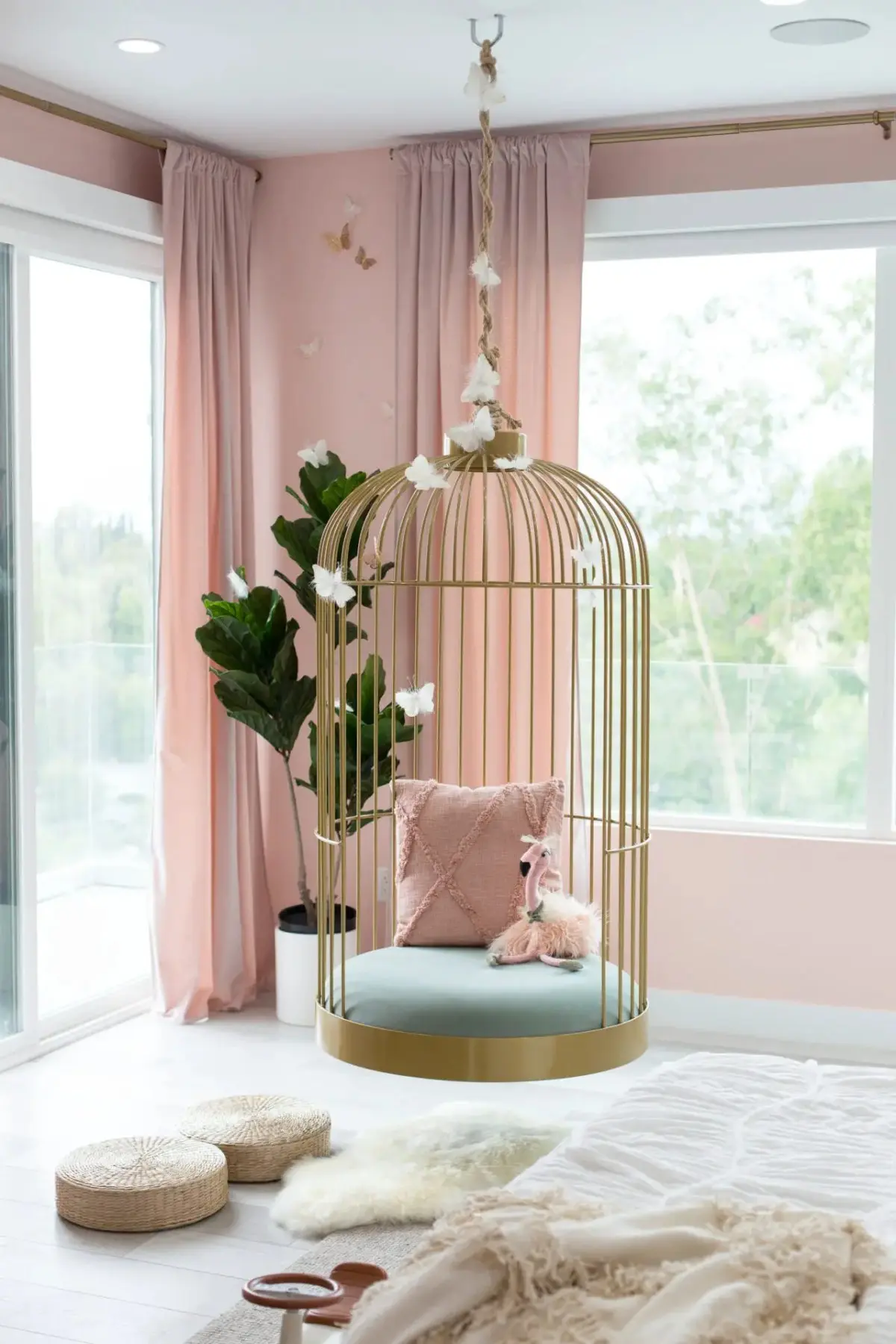 Room with a hanging birdcage chair decorated with pillows and a toy. Pink curtains, potted plant, and large windows in the background create an elegant atmosphere. Beige and white floor cushions in the foreground complete one of the 17 Pink Bedrooms Fit for a Princess.