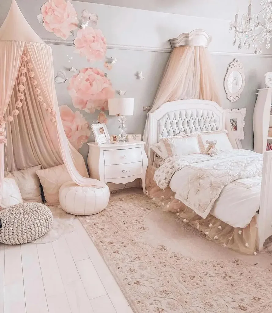 A cozy bedroom with a white and pink color scheme, featuring a bed with a tufted headboard, a canopy, a nightstand, and floral wall decorations. A play tent and plush seating are in the corner—one of 17 pink bedrooms fit for a princess.