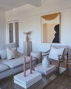 A modern living room with white sofas, wooden chairs, minimalist décor items, and a large abstract painting in warm tones.