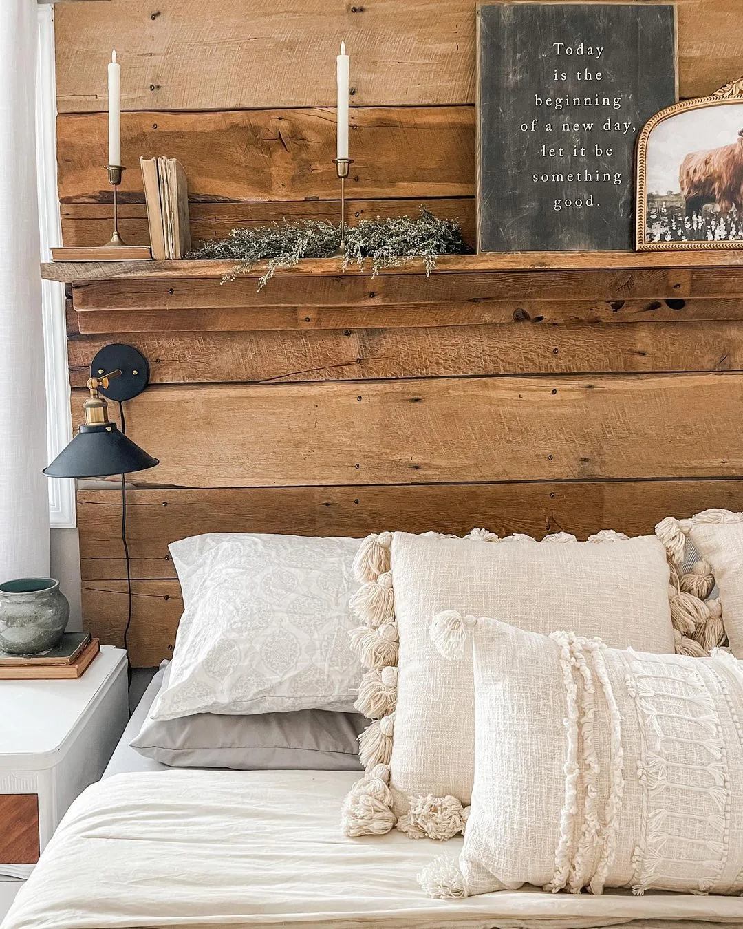 Cozy bedroom corner exuding farmhouse elegance with a white bed adorned with textured pillows, a rustic wooden headboard with shelves holding books, candles, and a decorative sign above.