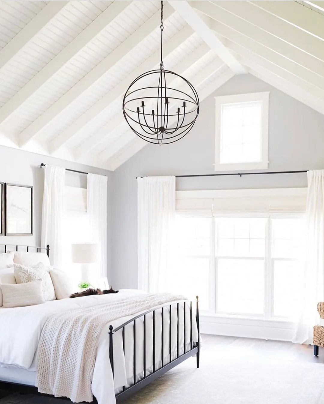 A bright, elegant bedroom featuring a vaulted ceiling with exposed beams, a large bed with a farmhouse-style black frame, white bedding, and a circular chandelier.