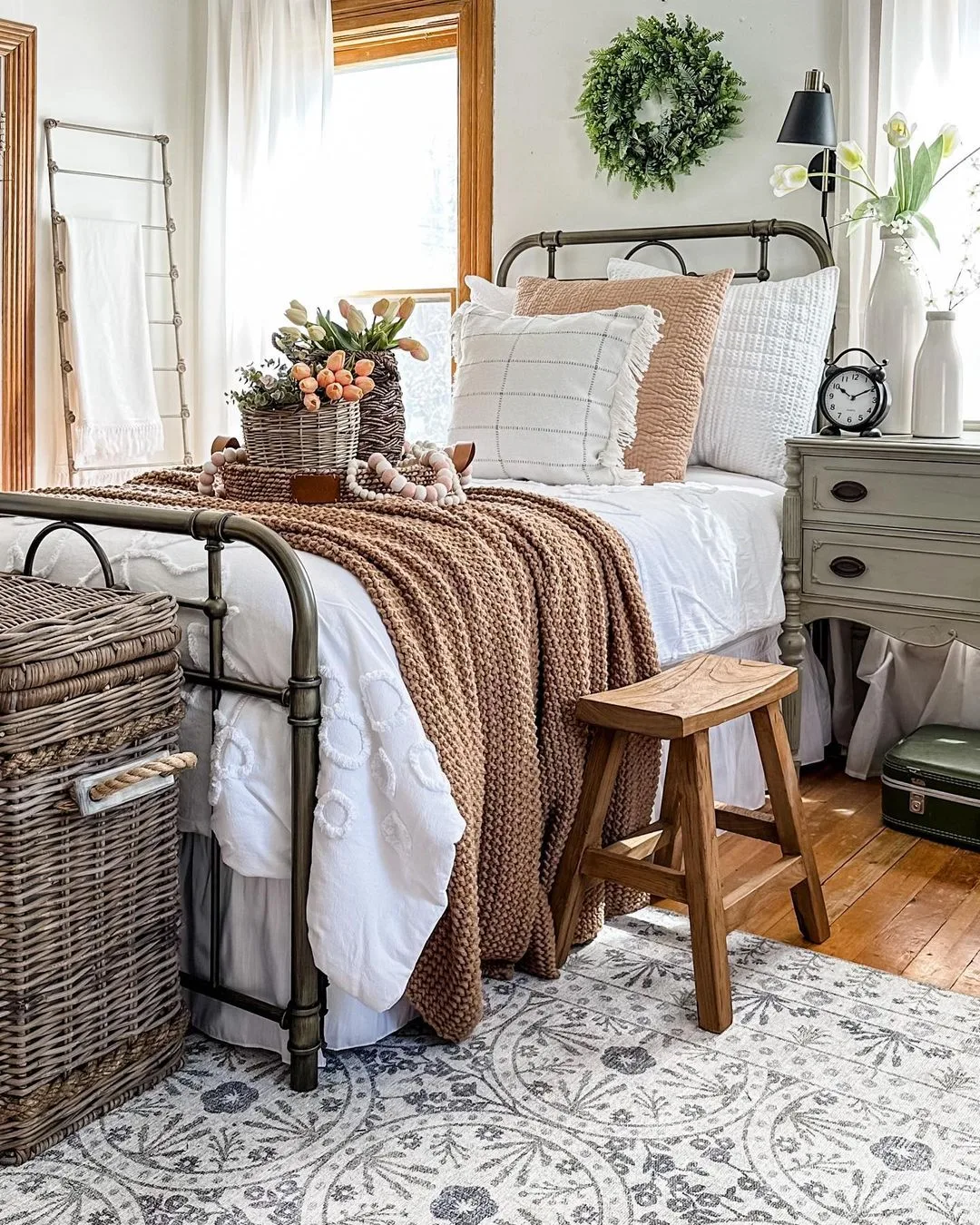 A neatly made bed with white and brown bedding, a wooden bench at its end, wicker baskets on the floor, and a decorated nightstand with a clock, lamp, and vase of flowers. A wreath hangs above the bed, epitomizing farmhouse elegance meets comfort in your personal bedroom retreat.