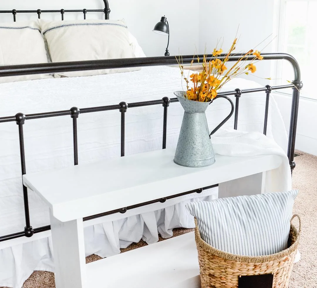 A bright bedroom with a black metal bed, white bench at the foot adorned with a vase of yellow flowers, beside an elegant wicker basket.