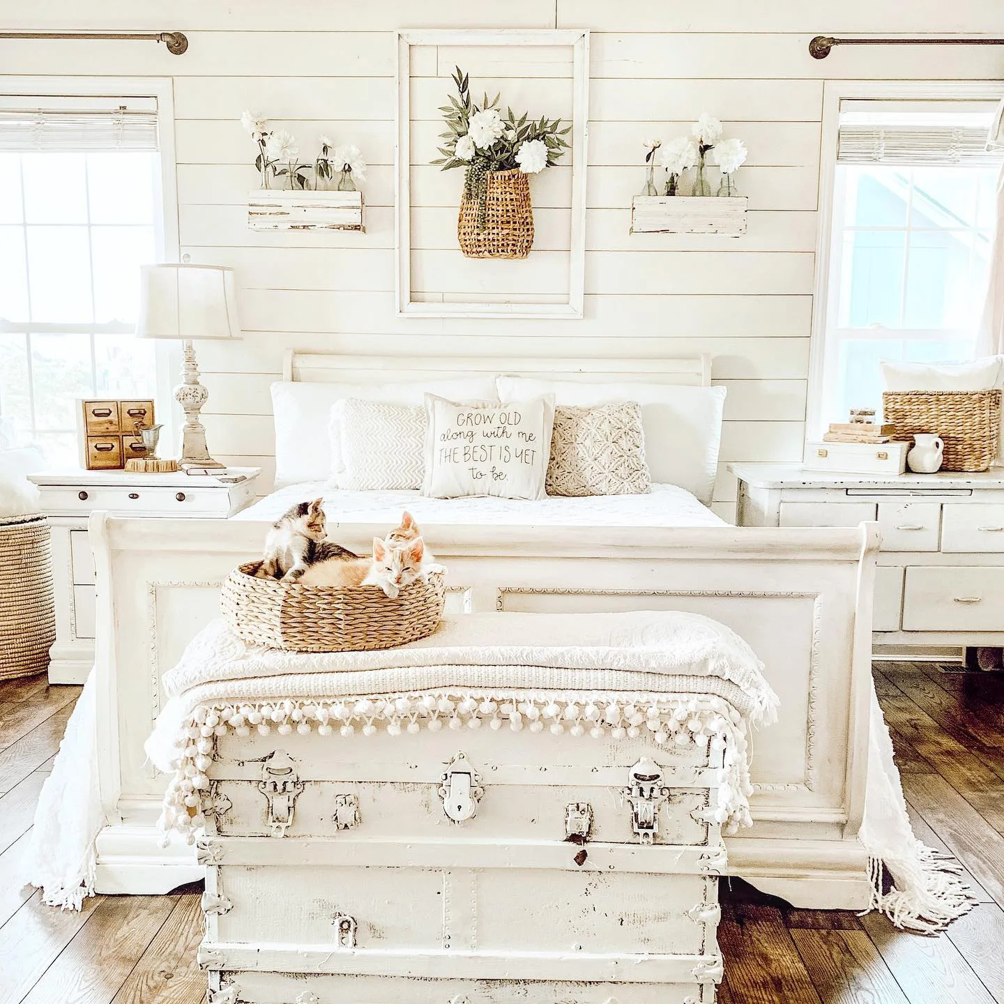 A bright, cozy bedroom with a vintage white bed, decorative pillows, and two cats resting in a farmhouse elegance wicker basket.