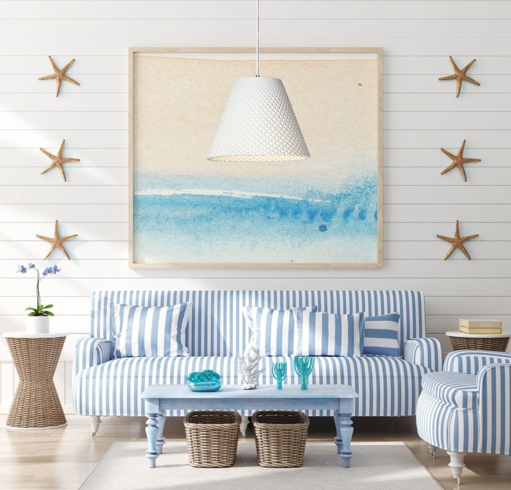 A coastal-inspired living room with a striped blue and white sofa, two wicker baskets, and a hanging pendant lamp. Coastal art and starfish decorations adorn the walls.