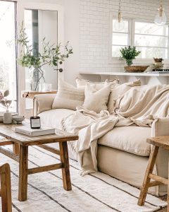 A cozy living room featuring a beige sofa with scattered pillows and a minimalist throw, accompanied by wooden stools and a small plant, with white tiled walls and pendant lights.