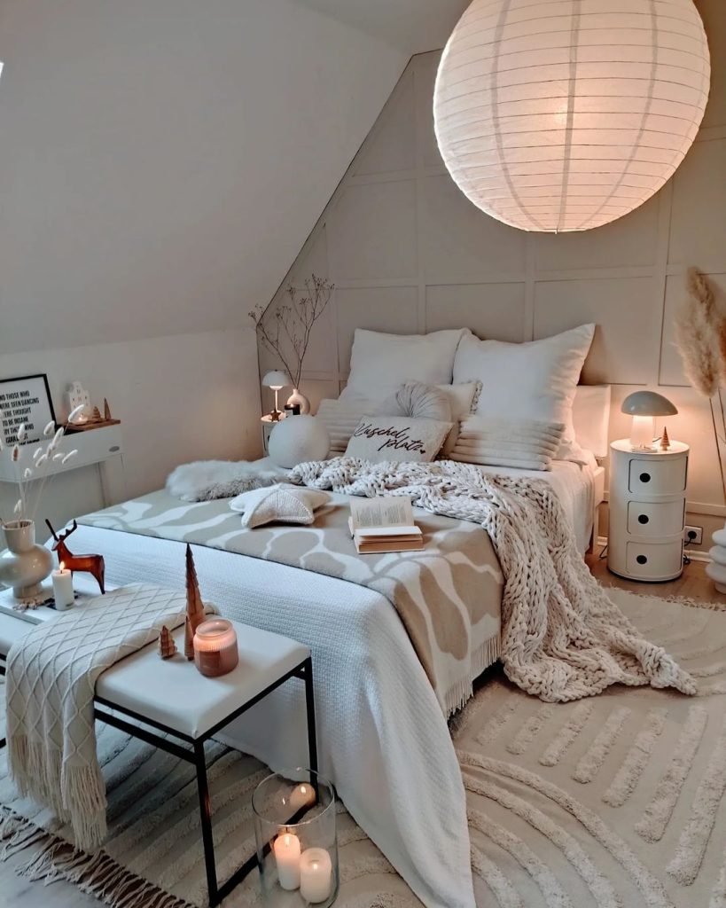 A cozy bedroom with a beige and white color scheme exudes boho-chic charm, featuring a neatly made bed adorned with multiple pillows, a large lantern-style lamp, side tables, candles, and decorative plants.