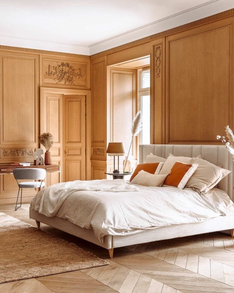Elegant Boho-chic bedroom with wooden paneling, a double bed with white and orange bedding, a desk area, and herringbone flooring.