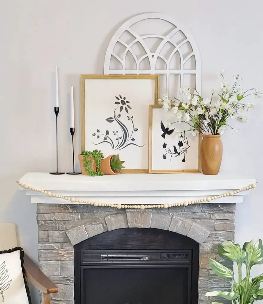 A tastefully decorated fireplace mantel featuring framed artwork, candlesticks, and a vase with fresh spring flowers, set against a stone surround.