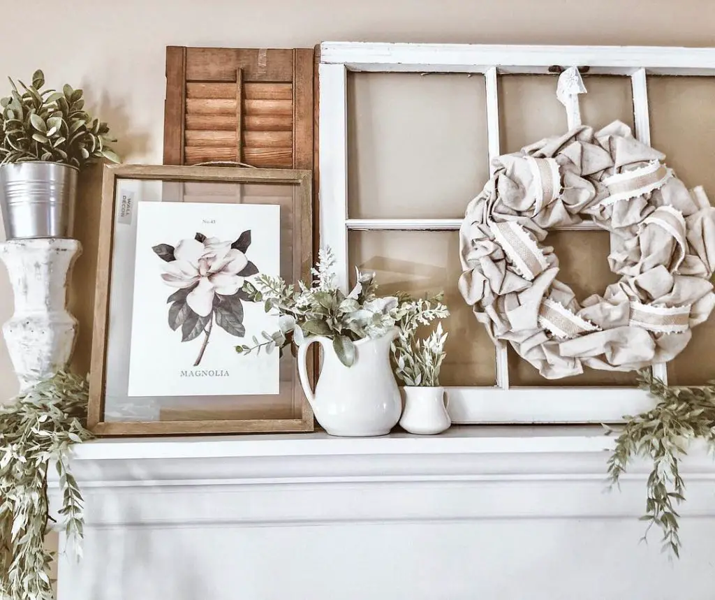 A rustic shelf displaying a vintage magnolia picture, a window frame wreath, a vase with flowers, and greenery, all in neutral tones.