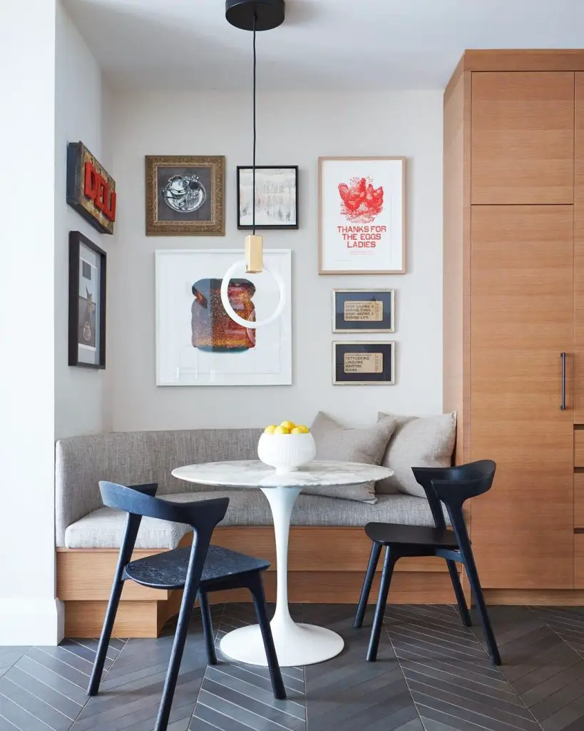 A cozy breakfast nook featuring inspiring banquette seating, a round white table, two black chairs, and a wall adorned with various framed artworks.
