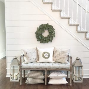 A cozy entryway with a farmhouse bench adorned with decorative pillows, flanked by two large lanterns, under a staircase with a green wreath hanging on the wall.