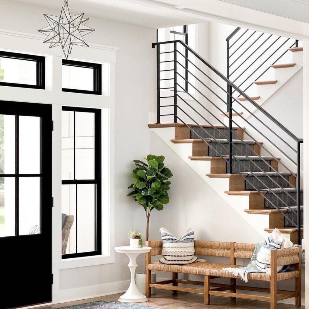 Modern interior with a wooden staircase, black railings, an entryway bench with cushions, and an indoor plant beside a large window. Decorative star-shaped pendant light overhead.