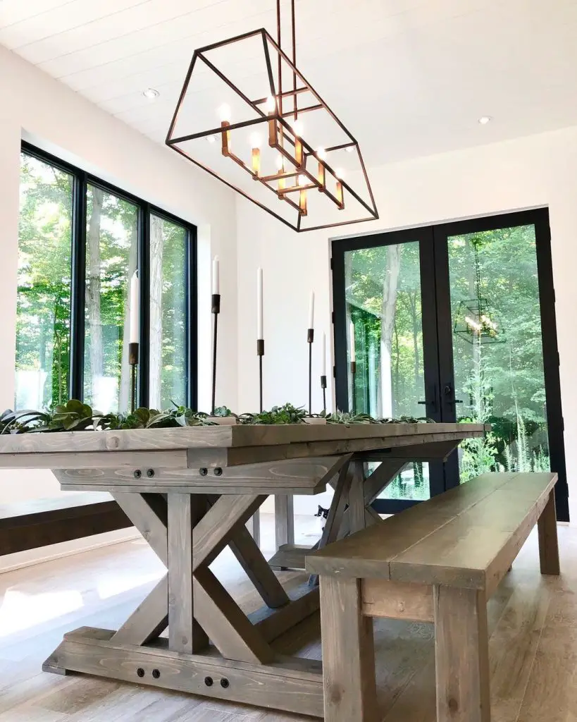 Modern dining room with a rustic farmhouse bench and table, geometric chandelier above, and large windows showing greenery outside.