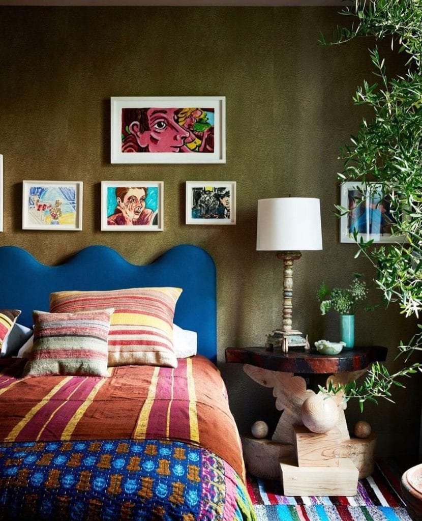 Cozy bedroom with an eclectic decor style featuring a vibrant blue headboard, multi-colored striped bedding, ornate wooden side table, and a collection of funky framed artwork above the bed.
