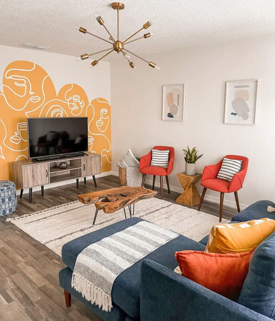 A modern living room with bold graphic wallpaper, two red chairs, a blue sofa, and a wooden TV stand. Decor includes abstract art and funky mid-century style chandelier.