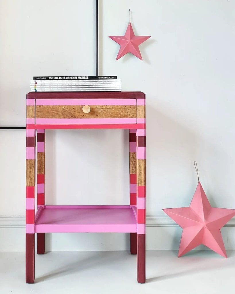 A pink and purple striped wooden nightstand with books on top, beside a hanging funky pink star and another star on the floor against a white wall.