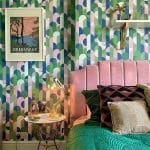 A stylish bedroom featuring a multicolored geometric wallpaper, pink tufted headboard, and decorative green bedding, with a framed poster and shelves on the wall.