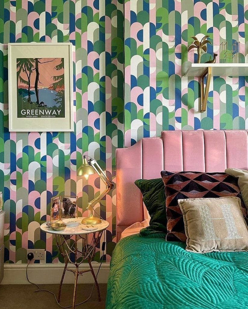 A stylish bedroom featuring a multicolored geometric wallpaper, pink tufted headboard, and decorative green bedding, with a framed poster and shelves on the wall.