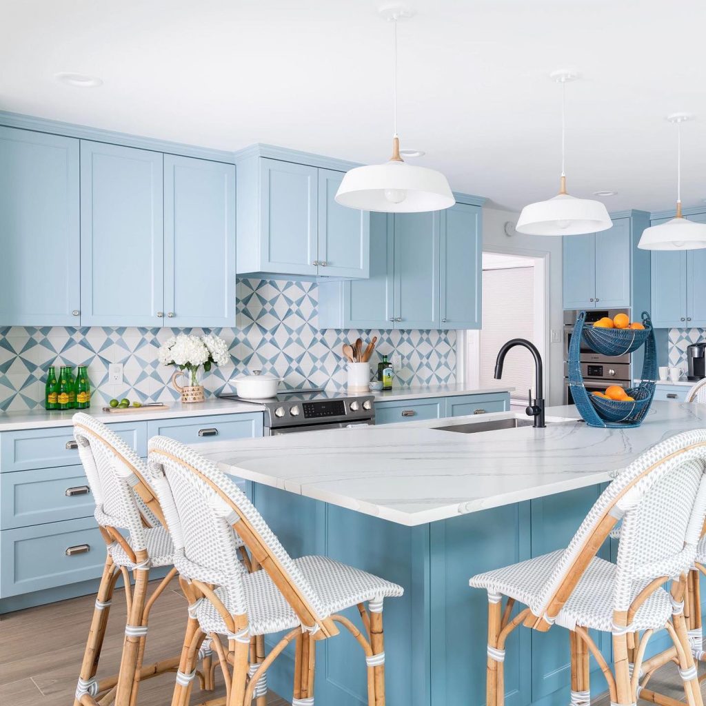 Modern kitchen with pale blue cabinets, white countertops, a central island, and hanging white lamps. Decor includes woven chairs and a trendy patterned backsplash.