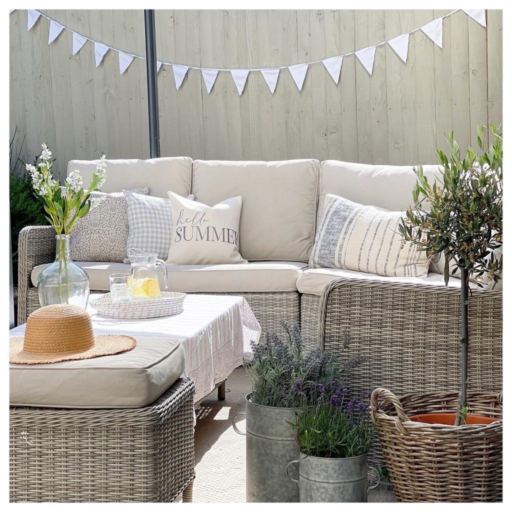 Cozy outdoor seating area with wicker furniture, decorative cushions in bright summer colors, and plants, featuring a 'hello summer' pillow, a straw hat on the table, and bunting overhead.