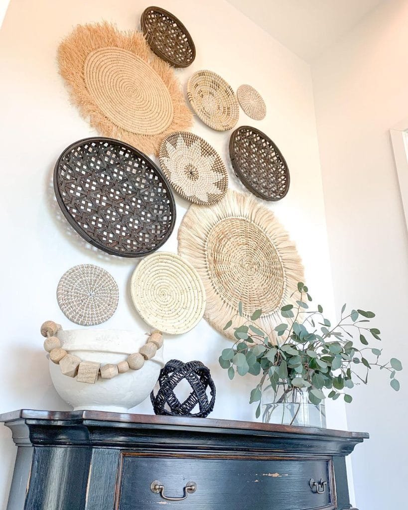 A variety of Bohemian wall baskets in different patterns and sizes displayed on a white wall above a dark wooden cabinet with decorative objects and a potted plant.
