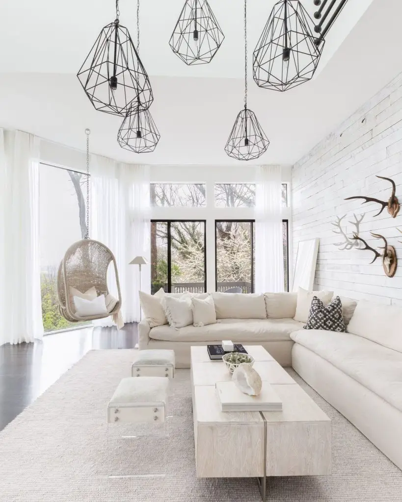 Brighten your trendy living room with a large white sofa, geometric pendant lights, hanging chair, and white brick wall against large windows overlooking trees.