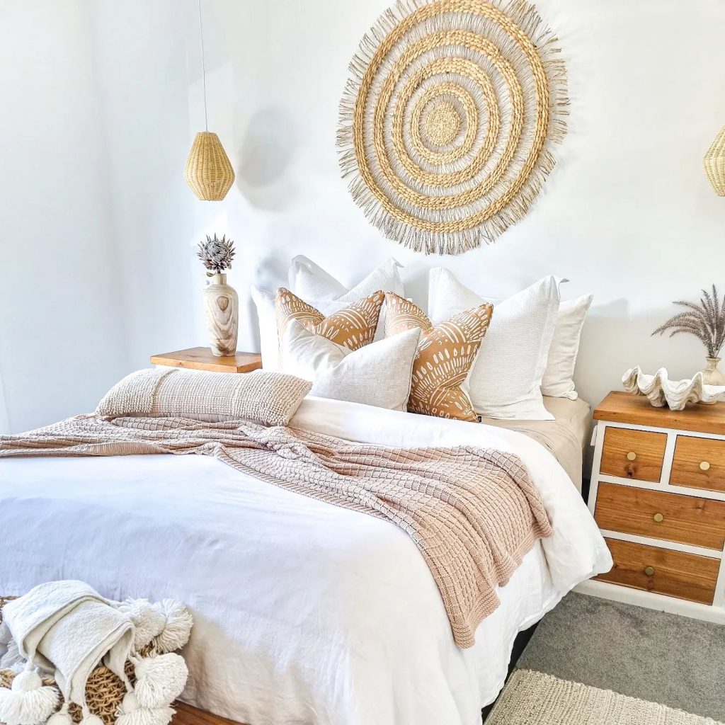 A cozy bedroom featuring a neatly made bed with white bedding, gold and white pillows, a round Bohemian woven wall decoration, and wooden bedside furniture. Light and airy ambiance.
