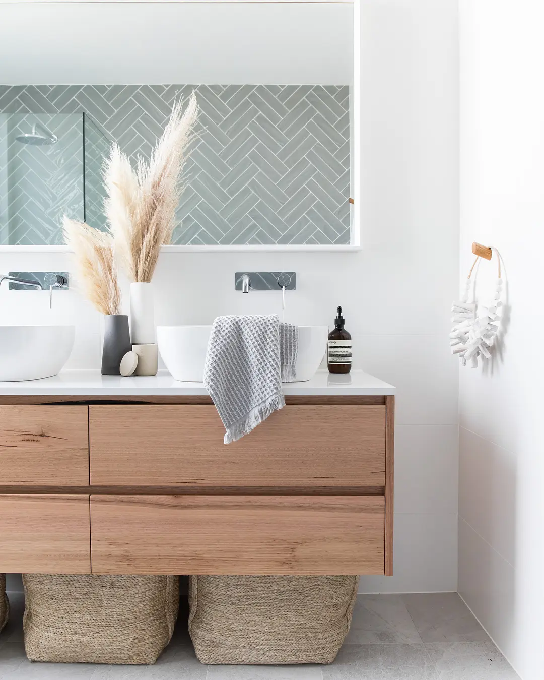 A modern Coastal Boho bathroom features a wooden vanity, double sinks, a large mirror, a herringbone-tiled backsplash, and decorative pampas grass in a vase. Woven baskets are stored under the vanity to complete this serene retreat.