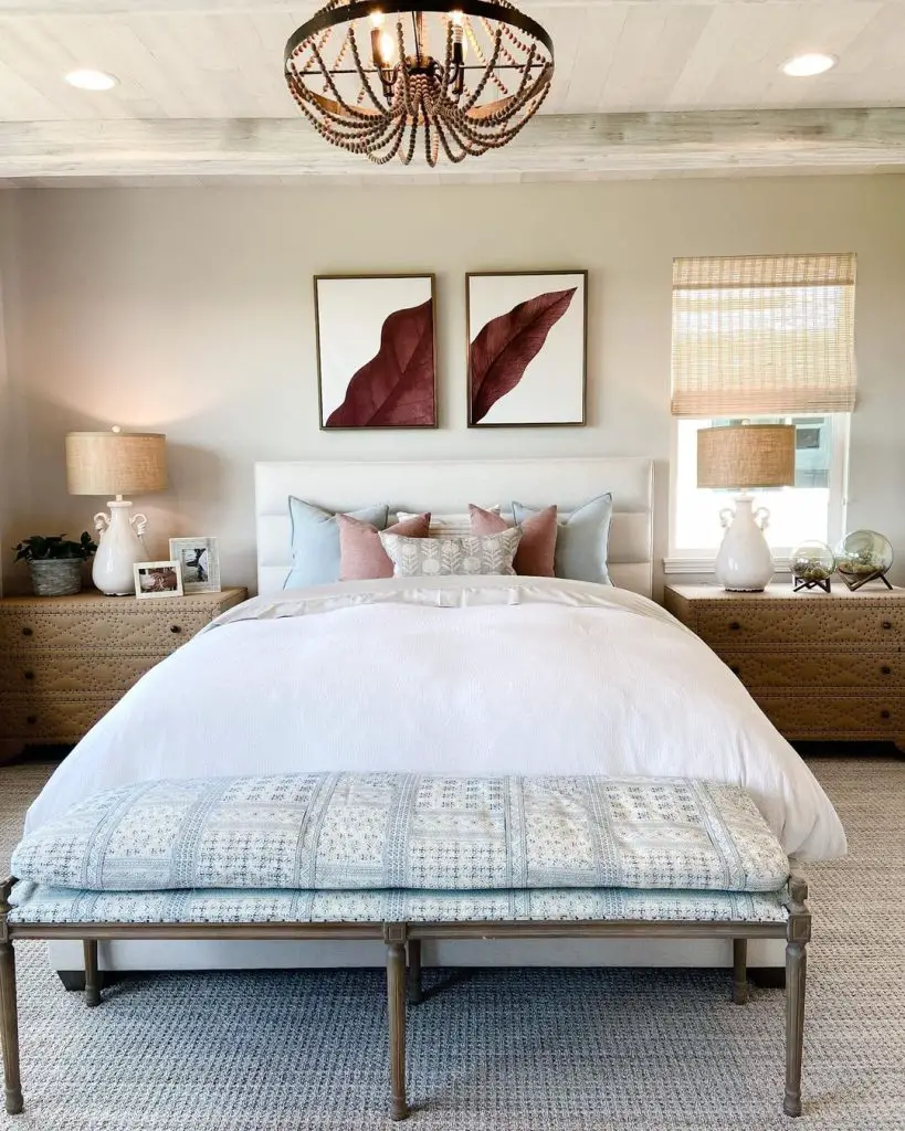 Elegantly designed bedroom with a king-sized bed, two coastal art pieces above, matching bedside tables with lamps, and a rustic chandelier.