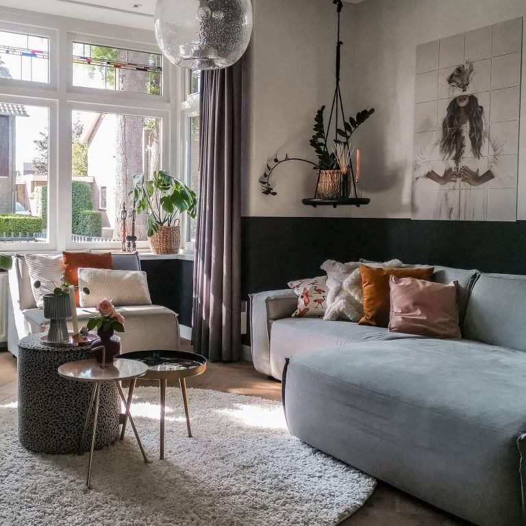 Modern living room with a gray sofa, eclectic textiles, round tables, and a large framed artwork above the sofa. Plants and large windows provide natural light.