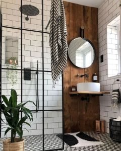 Modern bathroom with black and white tiled walls, a circular mirror, wooden accents, and a glass shower enclosure. A potted plant and striped towel add decorative touches.