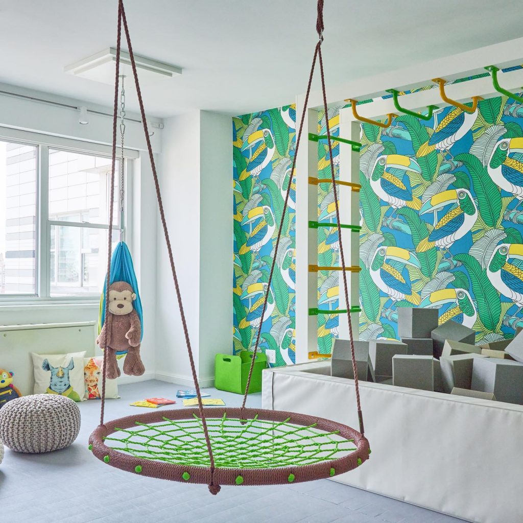 A playful children's playroom with a round swing, patterned curtains, toys, and a climbing ladder.