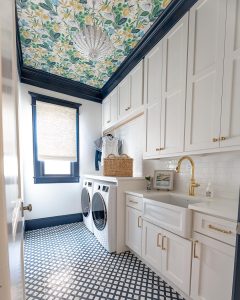 Laundry room with white cabinetry and appliances, patterned blue floor tiles, and a floral ceiling wallpaper. A dark blue window frame complements the decor. Learn how to maximize space in your long narrow laundry room.