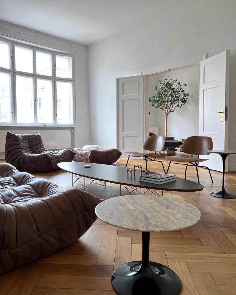 Modern living room demonstrating the perfect combination of minimalism and Mid-Century Modern style, featuring brown sofas, marble table, wooden chairs, and herringbone floor. A potted tree adds a touch of greenery.