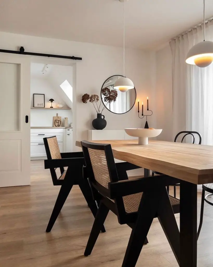 Modern dining room with wooden table and black chairs, pendant light above and a large circular mirror on the wall, leading to a bright kitchen area through a white doorway. Explore the 5 Essential Elements of a Minimalist Dining Room in this setup.