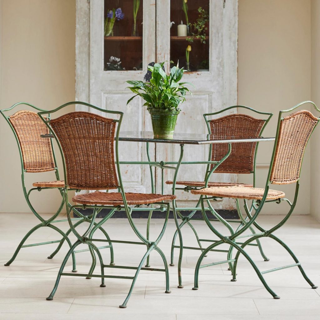 Four green metal and rattan chairs around a matching table, set in front of an ornate mirror in a room with a tiled floor.