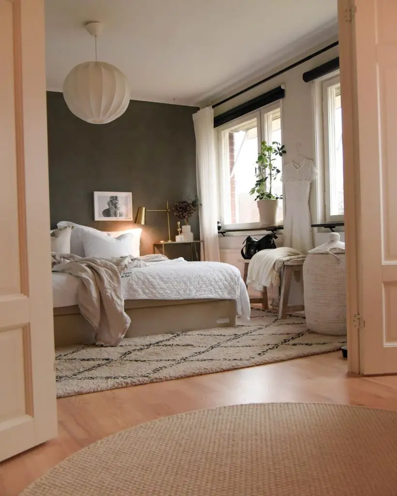 A cozy Scandinavian bedroom with soft lighting, featuring a gray accent wall, a white bed dressed in light linen, round rugs, and a potted plant by a window.